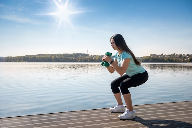 Hermosa joven se dedica a la gimnasia matutina con pesas en el lago