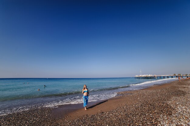 Hermosa joven delgada de pie en una playa de arena junto al mar. En traje de baño azul, vestido, falda chal de pareo. Turquía. vacaciones. Vacaciones. Luz de sol. Lugar para el texto