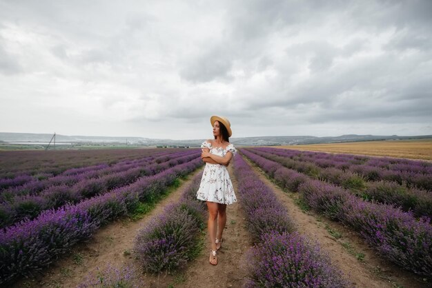 Una hermosa joven con un delicado vestido y un sombrero camina por un hermoso campo de lavanda y disfruta del aroma de las flores. Vacaciones y hermosa naturaleza.