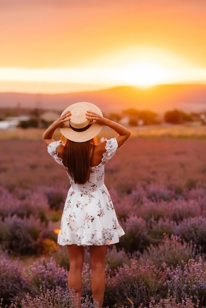 Una hermosa joven con un delicado vestido y un sombrero camina por un hermoso campo de lavanda y disfruta del aroma de las flores. Vacaciones y hermosa naturaleza.