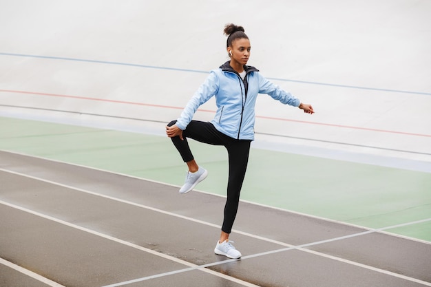 Hermosa joven deportista africana en forma con auriculares inalámbricos haciendo ejercicios de estiramiento en el estadio