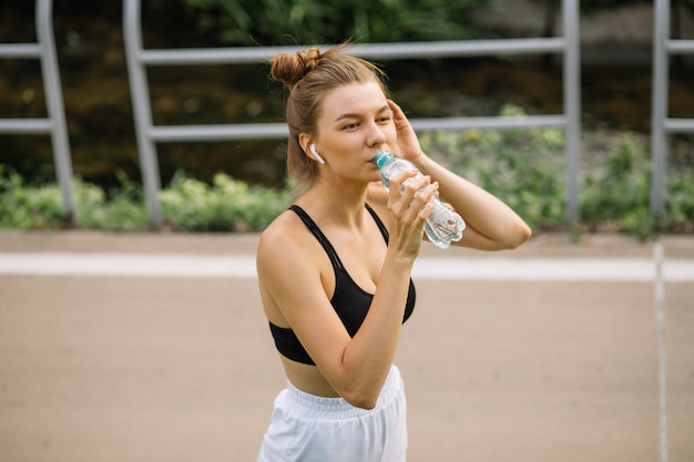 Hermosa joven deportista bebe agua de una botella de plástico, apaga su sed mientras hace deporte