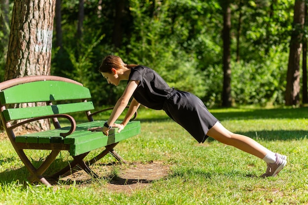 Hermosa joven deportista con camiseta negra, pantalones cortos negros y zapatillas rosadas calentando ejercicios de tríceps y inmersión en el pecho haciendo flexiones desde un banco entre los árboles antes de correr