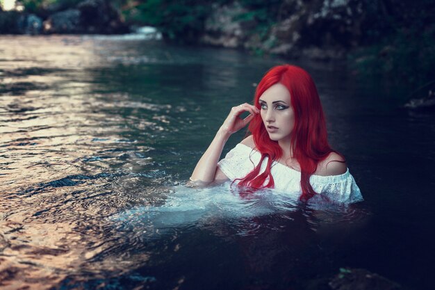 Hermosa joven descansando en el agua. Mujer joven con vestido blanco está sentada sobre la piedra en medio de un arroyo.