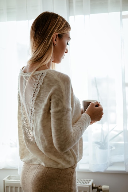 Hermosa joven disfrutando de la calma matutina y el café y mirando por la ventana.