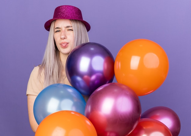 Hermosa joven disgustada con sombrero de fiesta de pie detrás de globos mostrando la lengua