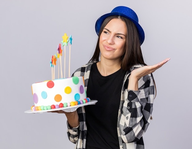 Hermosa joven disgustada con sombrero de fiesta sosteniendo la torta extendiendo la mano aislada en la pared blanca