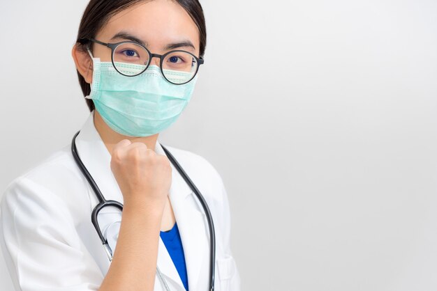 Hermosa joven doctora asiática con uniforme blanco y máscara protege el coronavirus, el médico levanta el puño para apoyar al paciente en la lucha contra la epidemia de enfermedades COVID 19, fondo gris del espacio de la copia