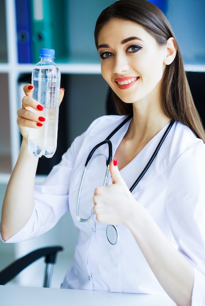 Hermosa y joven doctora en la sala de luz sosteniendo una botella de agua en sus manos