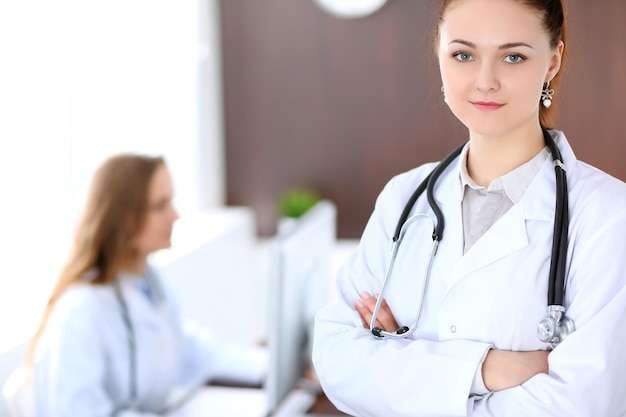 Hermosa joven doctora sonriente parada en un hospital con su colega en el fondo.