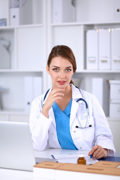 Hermosa joven doctora sonriente sentada en el escritorio y escribiendo