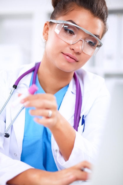 Hermosa joven doctora sonriente sentada en el escritorio y escribiendo