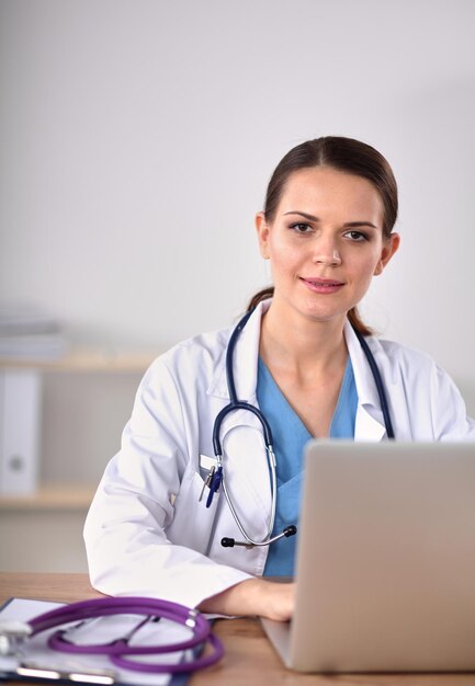 Hermosa joven doctora sonriente sentada en el escritorio y escribiendo