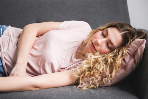 Hermosa joven durmiendo en un sofá gris Chica con camisa rosa descansando en casa después del trabajo