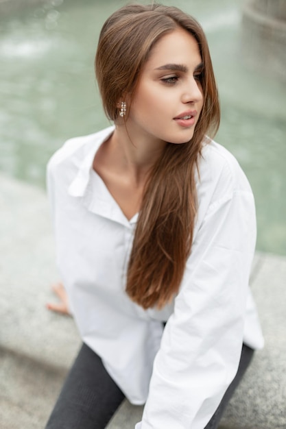 Hermosa joven y elegante chica de negocios con ropa de moda y una camisa blanca sentada cerca de una fuente en la ciudad