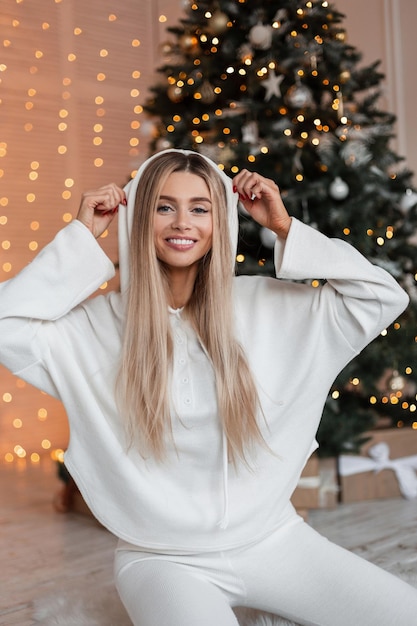 Hermosa joven elegante con una linda sonrisa vestida de blanco lleva una capucha y está sentada cerca del árbol de Navidad