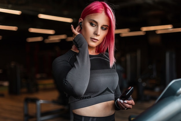 Hermosa joven elegante con maquillaje y cabello rosado poniéndose auriculares inalámbricos y comienza a correr en la caminadora en el gimnasio