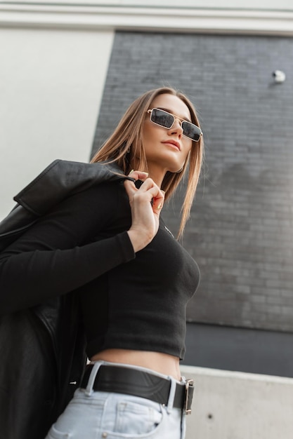 Hermosa joven y elegante modelo hipster con gafas de sol doradas vintage en ropa casual de moda con una camiseta negra con jeans y abrigo camina por la ciudad