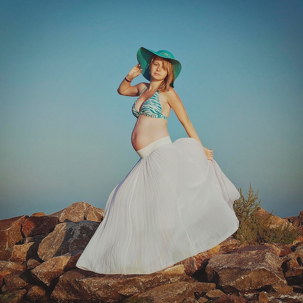 Hermosa joven y elegante mujer rubia embarazada con un sombrero azul camina por la playa, a la orilla del mar al atardecer