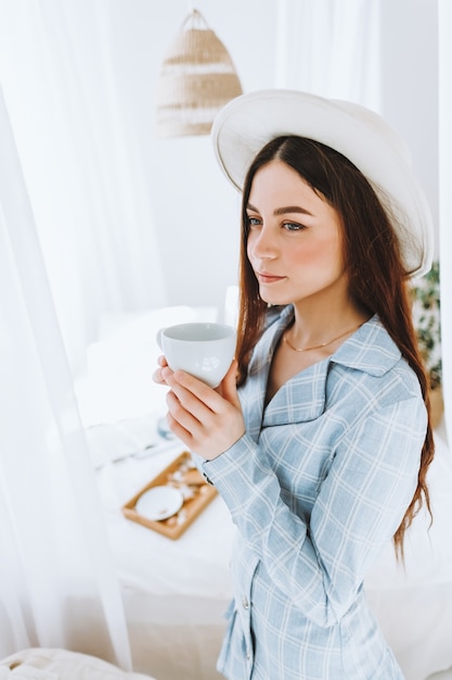 Hermosa joven elegante en traje y sombrero blanco de pie junto a la cama y beber café en la mañana soleada