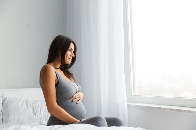 Hermosa joven embarazada en casa, sentada en la cama junto a la ventana