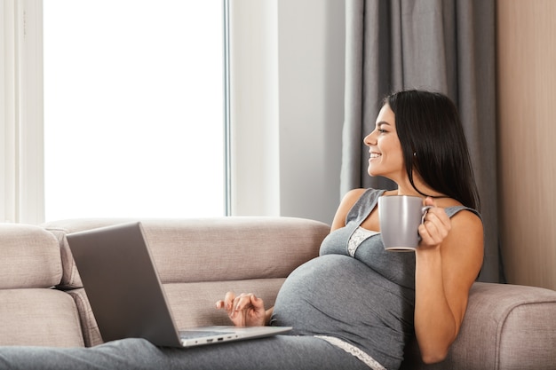 Hermosa joven embarazada en casa, sentada en un sofá, usando la computadora portátil, sosteniendo la taza