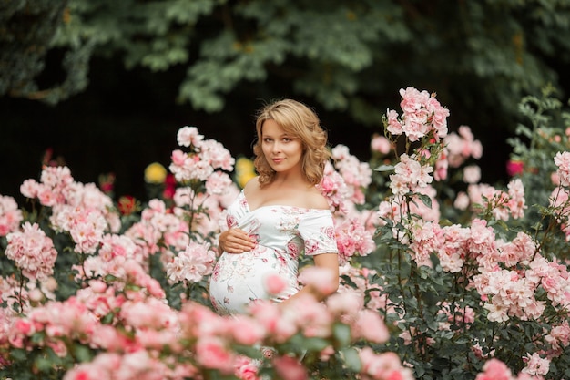 Una hermosa joven embarazada está caminando en un jardín de rosas. Retrato de una mujer embarazada con un vestido. El verano.