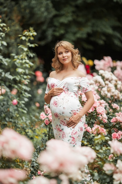 Una hermosa joven embarazada está caminando en un jardín de rosas. Retrato de una mujer embarazada con un vestido. El verano.