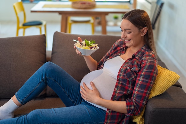 Foto hermosa joven embarazada feliz sentada y comiendo ensalada de frutas en el sofá en casa