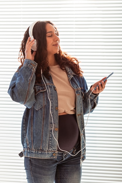 Hermosa joven embarazada pacífica escucha música clásica agradable con teléfonos inteligentes y auriculares. Concepto de actitud positiva antes del parto