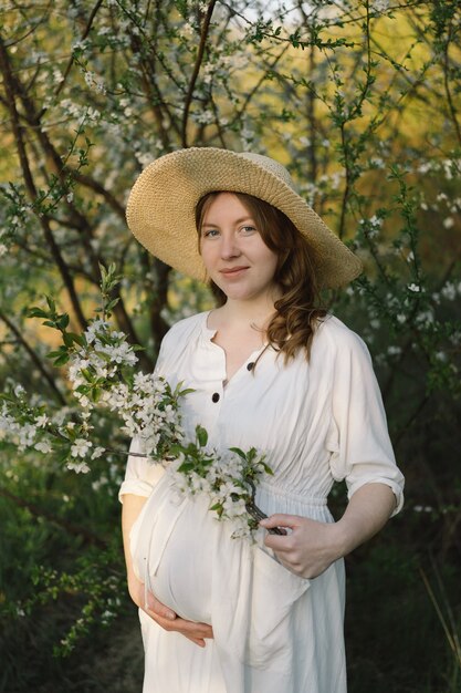 Una hermosa joven embarazada con un vestido blanco camina en el jardín de primavera