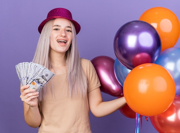 Hermosa joven emocionada con aparatos dentales y gorro de fiesta sosteniendo globos con dinero en efectivo aislado en la pared azul