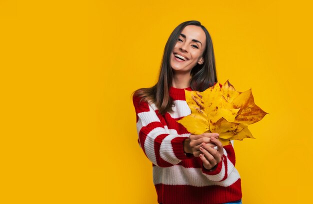 Una hermosa joven emocionada con un suéter a rayas muestra hojas amarillas en sus manos mientras se divierte y posa sobre fondo amarillo.