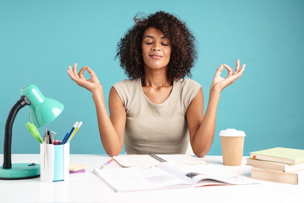 Hermosa joven empresaria africana sonriente vestida casualmente sentada en el escritorio aislado sobre la pared azul, meditando mientras estudia