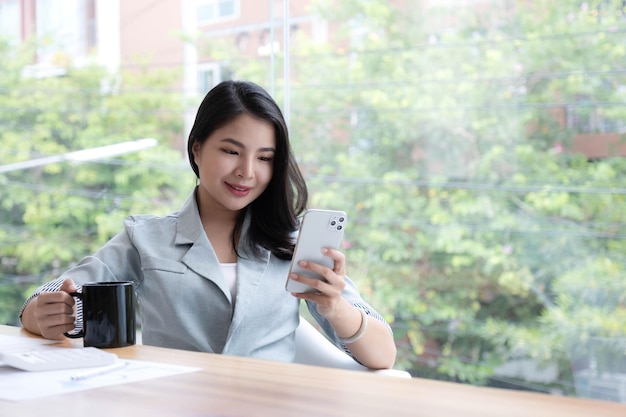 Hermosa joven empresaria asiática sonriendo sosteniendo una taza de café y un teléfono inteligente trabajando en la oficina