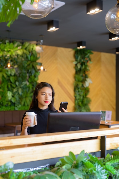 Hermosa joven empresaria asiática sosteniendo café mientras usa el teléfono en la cafetería.