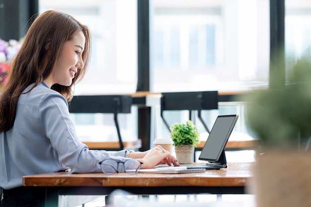 Hermosa joven empresaria asiática trabajando en tableta con felicidad mientras está sentado en la mesa en la sala de la oficina moderna.