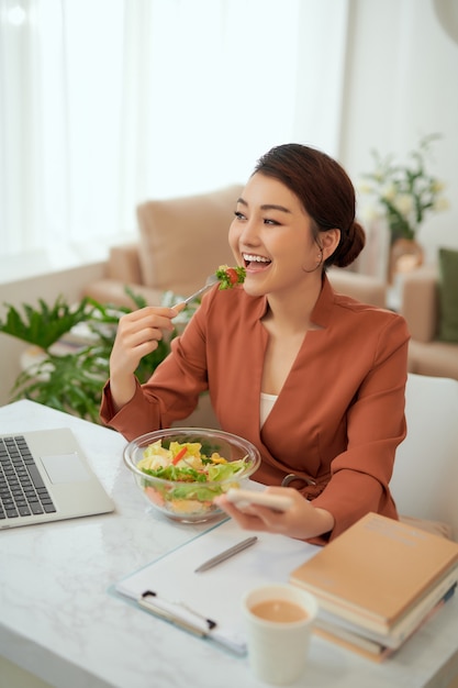 Hermosa joven empresaria en el lugar de trabajo, comiendo ensalada de verduras en un tazón,