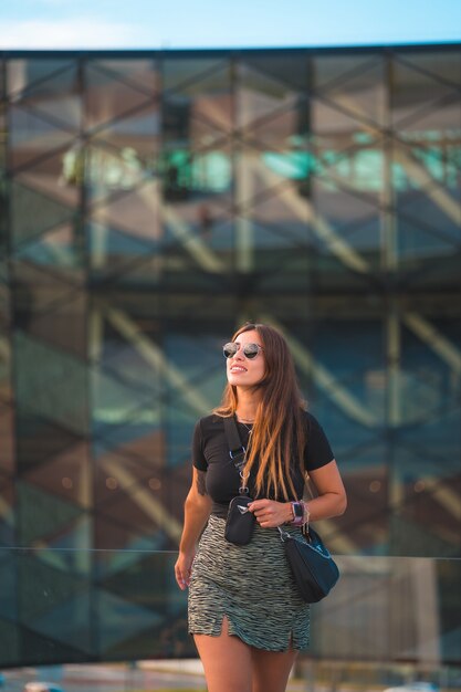 Hermosa joven empresaria morena caucásica fuera del fondo del edificio de oficinas de vidrio verde. Con gafas de sol y mirando, foto vertical