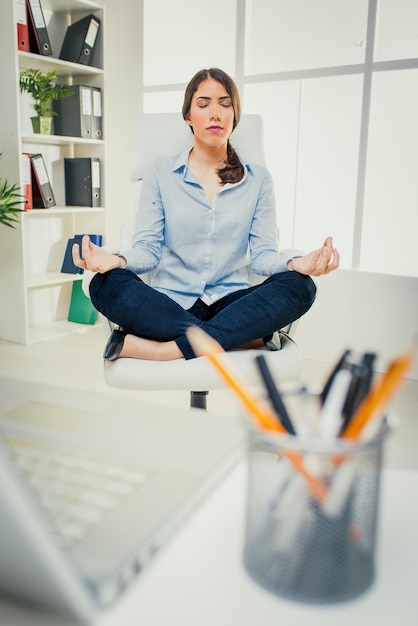 Foto hermosa joven empresaria practicando yoga en la oficina. ella se sienta en la postura del loto en una silla frente a su escritorio.