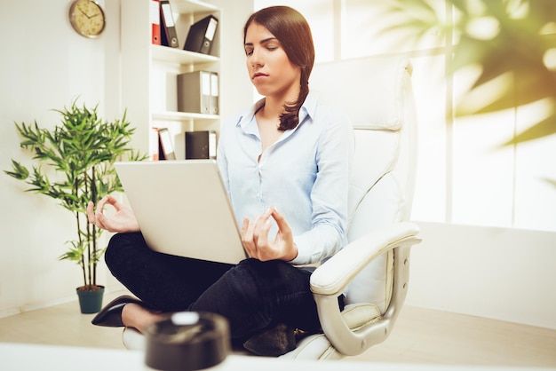 Hermosa joven empresaria sentada en posición de loto mientras meditaba en la oficina. Ella está practicando yoga y sosteniendo una computadora portátil en su regazo.