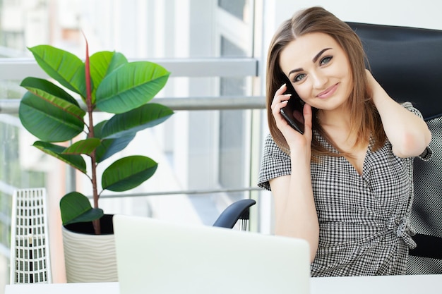 Hermosa joven empresaria trabajando en equipo portátil y hablando por teléfono.