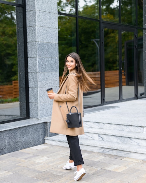 Hermosa joven empresaria vistiendo abrigo de otoño caminando al aire libre, sosteniendo la taza de café para llevar