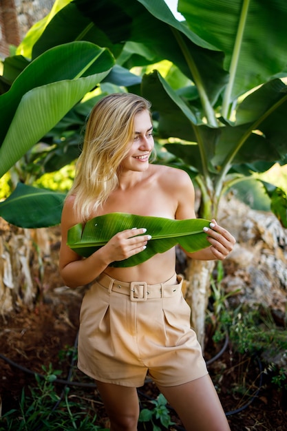 Una hermosa joven se encuentra en una hermosa pose cerca de un árbol de plátano, su niña cubre sus pechos con una hoja en una plantación de plátanos.