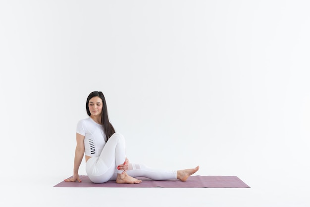 una hermosa joven se encuentra en la pose de Ardha Matsyendrasana sobre un fondo blanco