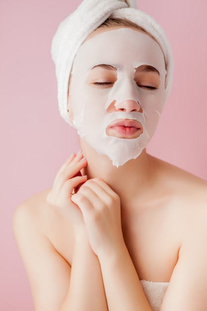 Hermosa joven está aplicando una máscara de tejido cosmético en una cara en un espacio rosa