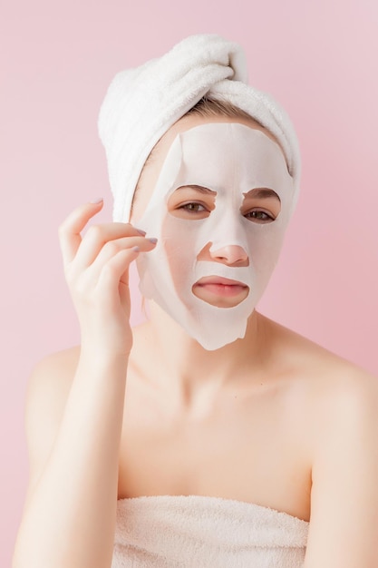 Hermosa joven está aplicando una máscara de tejido cosmético en una cara sobre un fondo rosa