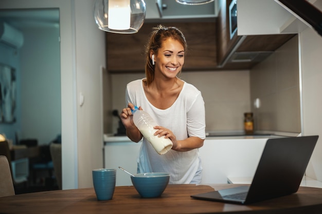 Hermosa joven está preparando su desayuno saludable en su cocina y usando una laptop para hacer un chat de video con alguien.