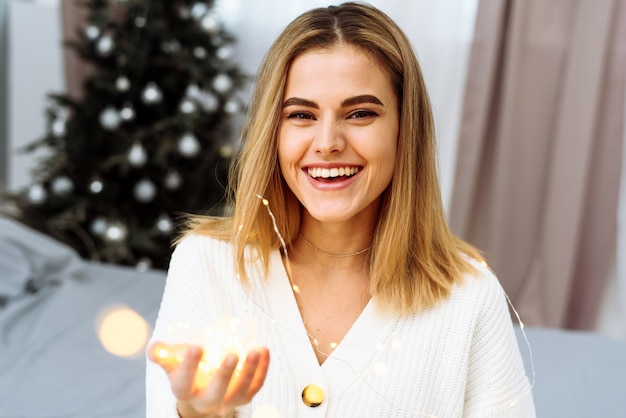 Una hermosa joven está sentada en una cama con el telón de fondo de un árbol de Navidad, sosteniendo una guirnalda en sus manos y sonriendo mirando a la cámara. Celebrando año nuevo y navidad en casa