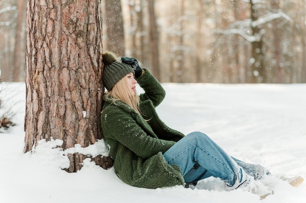 Hermosa joven está sentada cerca del árbol disfrutando del invierno al aire libre